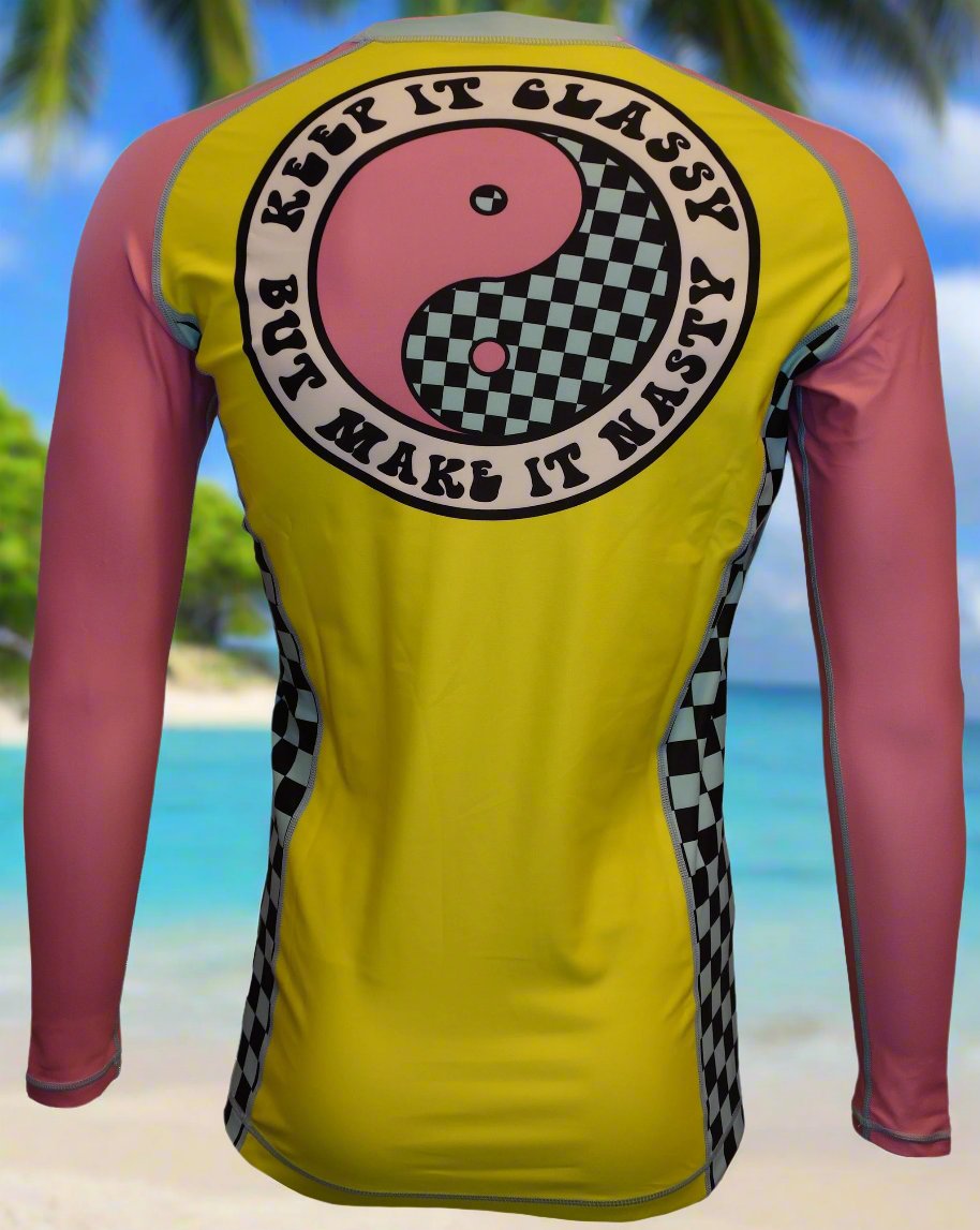 The Wave Hugger Classy and Nasty Rash Guard by Wave Hugger features yellow and pink long sleeves, a back yin-yang, checkered details, and “Keep it classy but make it nasty”—perfect surfwear for tropical beach vibes.